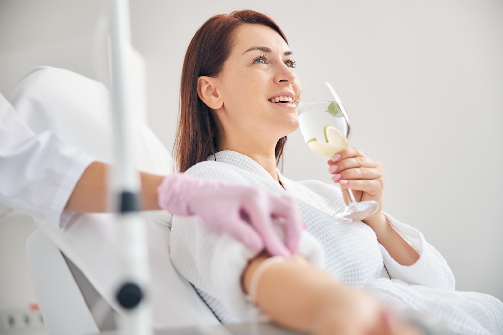 Attractive dark-haired woman smiling during the intravenous therapy Joyous female patient drinking a healthy beverage during a medical procedure in a beauty clinic